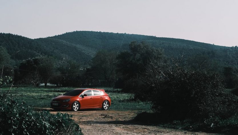 A vibrant red car parked amidst lush green hills and trees under a clear sky.